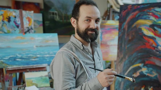 Portrait of handsome bearded man artist standing in studio next to colorful abstract picture and looking at camera wearing casual clothing and apron
