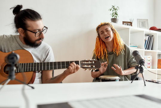 Live with the music. Man playing guitar and woman singing, playing rhythm with djembe drum while recording video blog or vlog. Couple of musicians making music at home