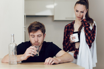 Drinking problem drunk man in a young family. A man drinks while sitting at a table in the kitchen, an upset woman stands next to a man and looks at him