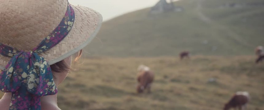 Young female farmer woman wear straw hat stops to look at her mountain cow herd on a grass meadow field pasture in autumn or fall evening light close up slow motion flare