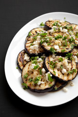 Homemade Grilled Eggplant with Feta and Herbs on a white plate on a black surface, side view.
