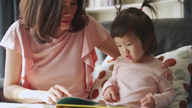 Asian beautiful young mother and her little daughter reading documentary book on sofa at home together. They pointing on the book looking it with happiness spending time together. Happy family concept