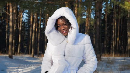 smiling African-American girl in white fur coat with hood poses against high pine trees in winter park closeup