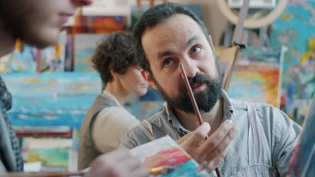 Experienced arts teacher is teaching male student who is painting picture in class while diligent young woman is working with portrait in background