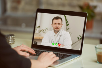 Fototapeta na wymiar A laptop screen view over a woman's shoulder. A woman at an online doctor's appointment. A girl is discussing her health with a therapist in a white lab coat on a video call.