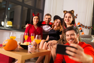 friendship, holiday and people concept - group of happy smiling friends in halloween costumes of vampire, devil, witch and cheetah taking selfie by smartphone at home party at night
