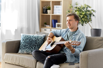 leisure, music and people concept - young man or musician with guitar, earphones and smartphone sitting on sofa at home