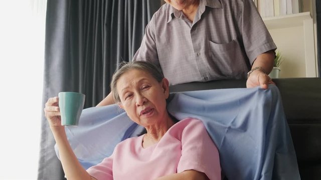 Mature Old Man Taking Care Woman While Drinking Hot Coffee Or Tea Sitting On Sofa In Living Room. Elderly Husband Give Blanket Cover For Wife And Hug From Behind. Happy Senior Retirement Lifestyle.