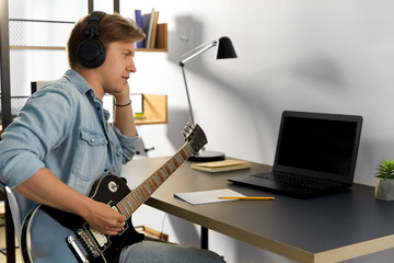 leisure, music and people concept - young man or musician in headphones with laptop computer playing bass guitar sitting at table at home