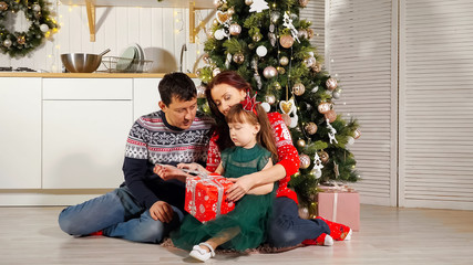 Obraz na płótnie Canvas girl child in green dress unpacks New Year gift box sitting at Christmas tree with parents and getting kisses