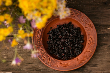 A round plate of red clay with blackberries stands on an old wooden table next to a bouquet of wildflowers; blurred background