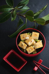 Roasted chunks of tofu cheese in a red bowl, vertical shot over black stone background with green ficus tree, elevated view