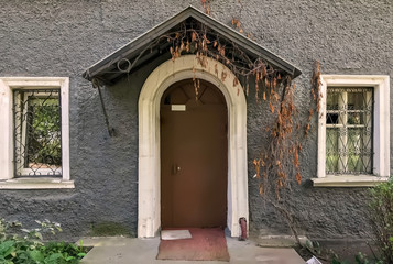 Fragment facade of an old house with dark brown door surrounded by gray stucco wall. White arched opening around door and triangular canopy over. Porch of residential building with Windows and plants