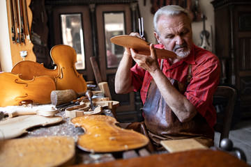 Arts and crafts. Senior carpenter craftsman checking sound quality of wood material in his old-fashion carpenter's workshop. In background hardwood violin parts and tools.