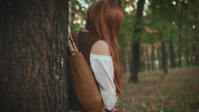 Medieval red-haired woman stands in the forest under a tree. The girl holds a wooden bow in her hands. Fantasy image of Lady hunter. Vintage warrior archer. Leather pants, white shirt, jacket