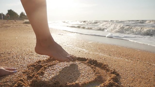 Close-up of woman's leg draws on the sand on the beach near the sea, ocean