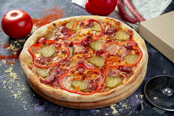 Oven-baked Italian pizza with pickled cucumber, bell pepper, bacon and smoked sausages on a dark background