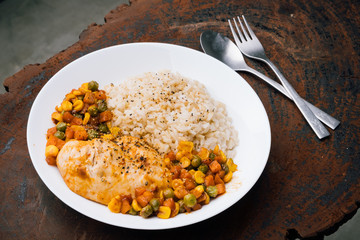 a dish of homemade roasted chicken breast and dice carrots, beans, corns with brown rice on dark red wooden table and cutlery