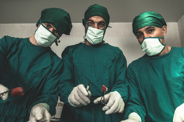 Low angle view portrait group of surgeons holding medical equipment and looking at camera during surgical operation in operating room. Medical team working to save patient life in hospital background