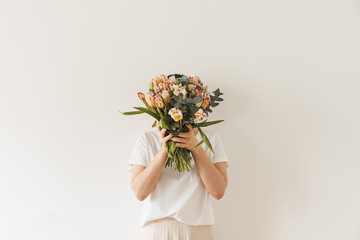 Young pretty woman in white blouse holding tulip flowers bouquet in hands against white wall....