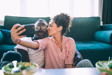 Multiethnic couple taking selfie with smartphone while kissing at home