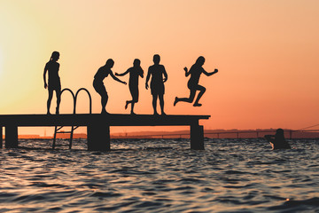 Silhouettes of people jumping from the pier into the sea at sunset.