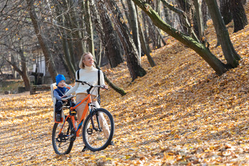 Fototapeta premium Happy mother leads a bicycle with a child strapped in the back in the autumn park.