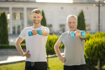 Young male and grey-haired man holding dumbbell in one hand, another hand on hip