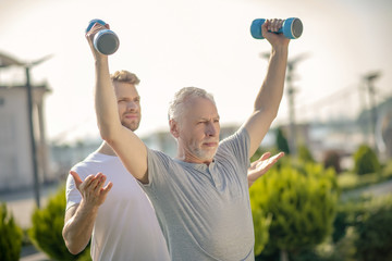 Grey-haired man raising hands with dumbbells, bearded instructor helping him