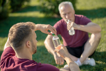 Obraz premium Bearded male opening bottle of water, grey-haired man drinking water, both sitting on grass