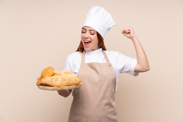 Redhead teenager girl in chef uniform. Female baker holding a table with several breads celebrating a victory
