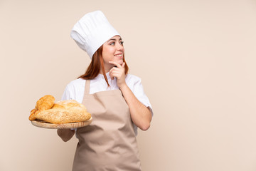 Redhead teenager girl in chef uniform. Female baker holding a table with several breads thinking an idea and looking side