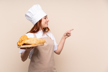 Redhead teenager girl in chef uniform. Female baker holding a table with several breads pointing to the side to present a product