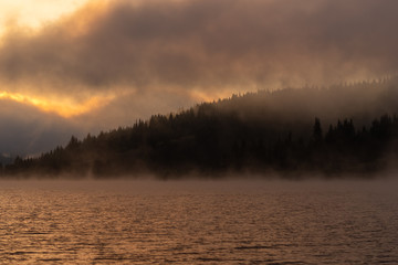 Dramatic view of a natural landscape in a mountainous area with a quiet lake and a coniferous forest shrouded in fog with the sun rising behind the clouds in the early hours of the morning.