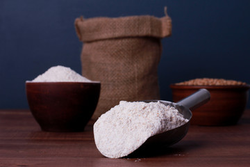 Metal scoop and bowl with wholegrain flour powder on brown wooden table against sack of wheat...