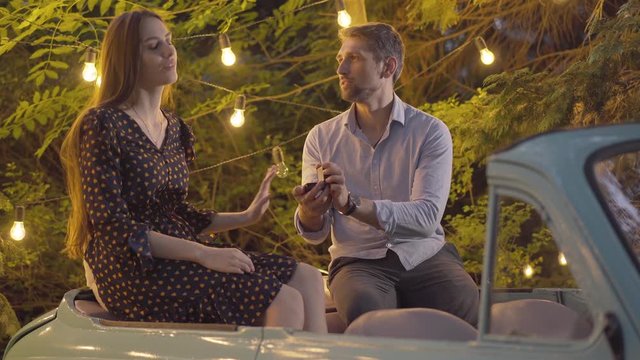 Young beautiful woman rejecting proposal of loving man. Portrait of Caucasian girlfriend refusing marriage. Shocked boyfriend with wedding ring sitting with lover in retro car on summer evening.