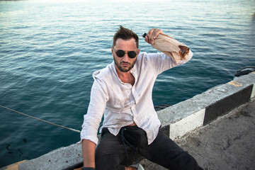 Handsome man with beard relaxing on the street  parapet, smoking ciggarette, drinking and looking to the view of the sea