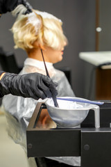 Hairdresser in black gloves dyes hair of young woman in white color in hairdress salon. Blured background