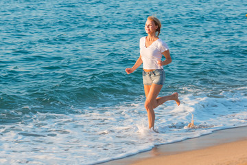 Smiling adult woman in white T-shirt is jogging on the beach near the ocean.