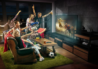Group of friends watching TV, football match, sport together. Emotional men and women cheering for...