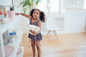 Black little girl with strainer standing near kitchen shelf