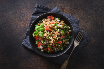 Salad of whole grain cereal spelt with seasonal vegetables, tomato, cucumber in bowl on dark background. Top view.