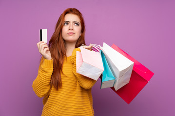 Redhead teenager girl over isolated purple background holding shopping bags and a credit card and thinking