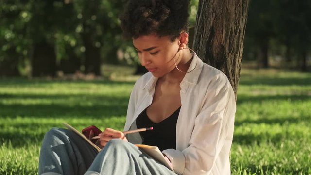 A pleased young african american woman is drawing sitting on the grass in the park