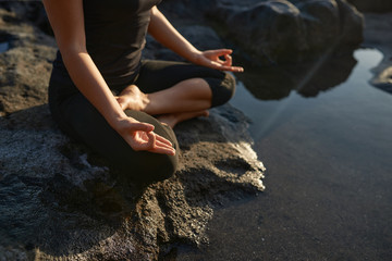 Woman practicing yoga and meditation in lotus position on black stones near the sea.