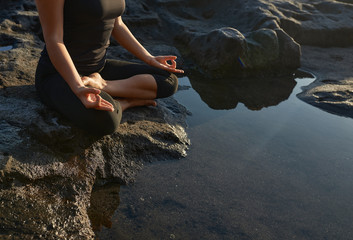 Woman practicing yoga and meditation in lotus position on black stones near the sea.