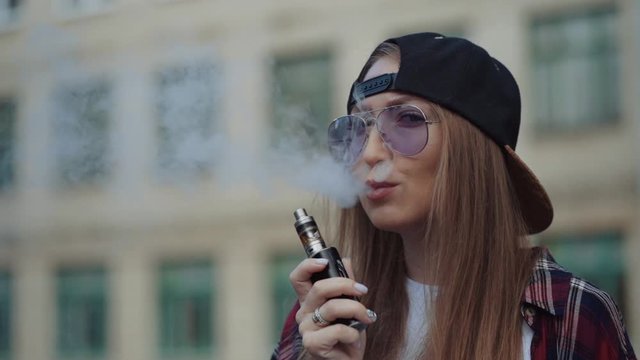 Close up pretty young hipster woman in black cap vape ecig. Girl vape popular ecig gadget, vaping device.Happy brunette vaper girl with e-cig. Portrait of smoker female model with electronic cigarette