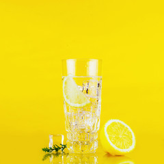 A glass of gin and tonic with lemon and ice cubes on yellow background