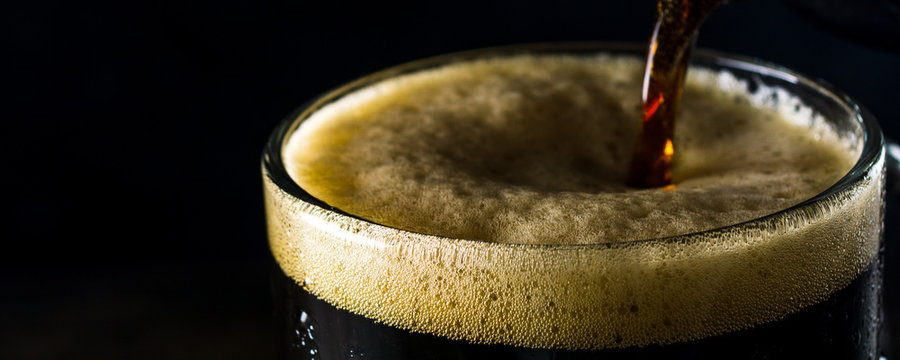 Porter beer pours from a bottle into a mug on a dark background. Panoramic frame, close up and copy space for text