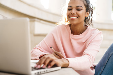 Image of cheerful african american student girl doing homework with laptop - Powered by Adobe